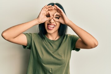Young hispanic girl wearing casual t shirt doing ok gesture like binoculars sticking tongue out, eyes looking through fingers. crazy expression.