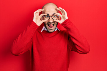 Young bald man wearing casual clothes and glasses doing ok gesture like binoculars sticking tongue out, eyes looking through fingers. crazy expression.