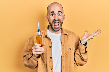 Young bald man holding olive oil can celebrating achievement with happy smile and winner expression with raised hand