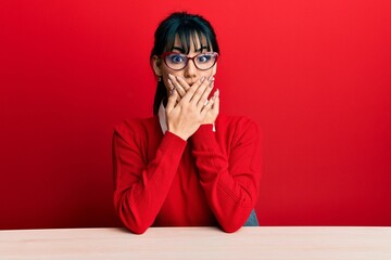 Young brunette woman with bangs wearing glasses sitting on the table shocked covering mouth with...