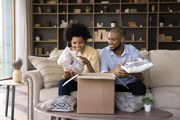 Happy young mixed race husband and wife unpacking cardboard box, producing warm Christmas clothes,...