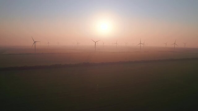 Windmills with rotating wings at sunset or sunrise. Technology for wind energy, renewable energy source, earth care. Wind power station, drone video, moving toward the horizon, 4k