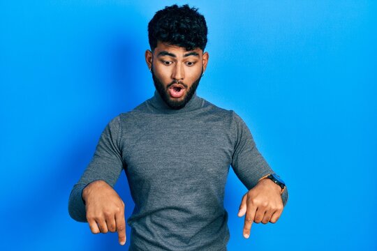 Arab man with beard wearing turtleneck sweater pointing down with fingers showing advertisement, surprised face and open mouth