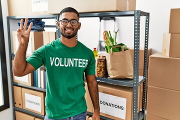 Young indian man volunteer holding donations box showing and pointing up with fingers number three while smiling confident and happy.
