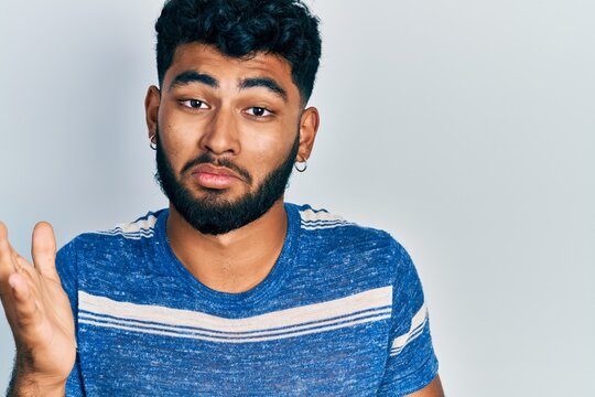 Arab man with beard wearing casual striped t shirt clueless and confused expression with arms and hands raised. doubt concept.