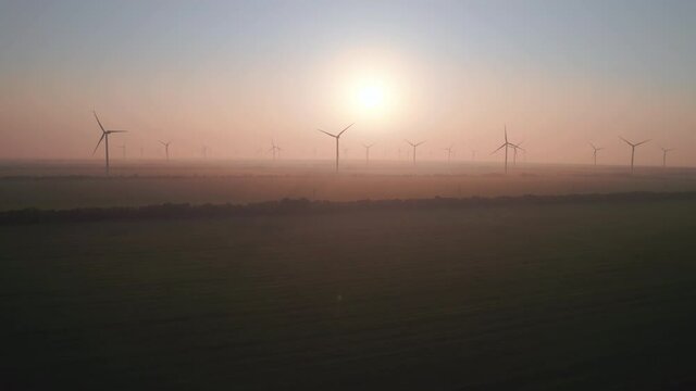 Windmills with rotating wings at sunset or sunrise. Technology for wind energy, renewable energy source, earth care. Wind power station, drone video, moving toward the horizon, 4k
