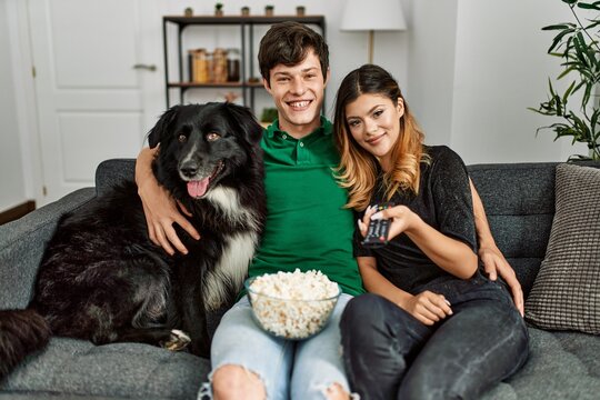 Young caucasian couple watching movie sitting on the sofa with dog at home.