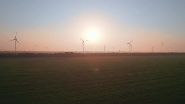 Windmills with rotating wings at sunset or sunrise. Technology for wind energy, renewable energy source, earth care. Wind power station, drone video, moving toward the horizon, 4k
