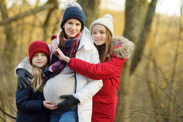 Obraz premium Young pregnant woman hugging her older daughters. Older siblings having fun with her pregnant mom outdoors. Mother and her kids spending time together.