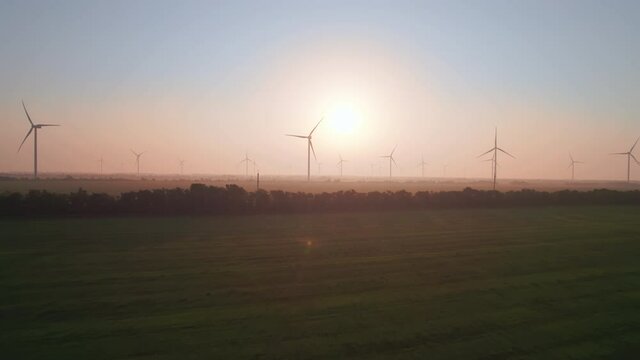 Windmills with rotating wings at sunset or sunrise. Technology for wind energy, renewable energy source, earth care. Wind power station, drone video, moving toward the horizon, 4k
