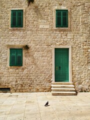 Fototapeta na wymiar Green door, three shuttered windows and lonely dove in the yard of the house built of shell-rock stones