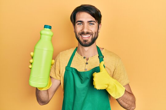 Young hispanic man wearing cleaner apron holding cleaning product smiling happy and positive, thumb up doing excellent and approval sign