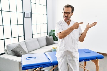 Middle age physiotherapy man working at home amazed and smiling to the camera while presenting with hand and pointing with finger.