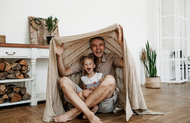 a happy father plays with his little daughter on the floor making a tent out of a blanket.