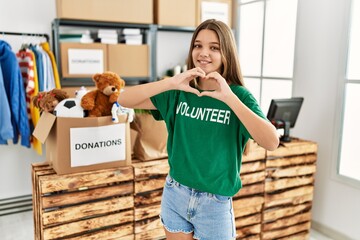 Adorable girl wearing volunteer uniform doing heart symbol with hands at charity center