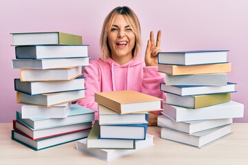 Young caucasian woman sitting on the table with books smiling with happy face winking at the camera doing victory sign with fingers. number two.