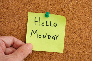 A man holding a sticky note that is pinned to a corkboard with the greeting Hello Monday on it