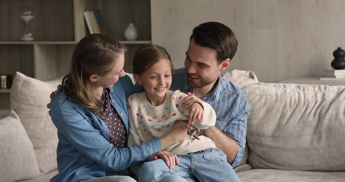 Overjoyed little girl cuddling with laughing mother father on sofa hold keys from new rented purchased apartment in hand. Excited family young parents preteen daughter get mortgage loan to buy a flat