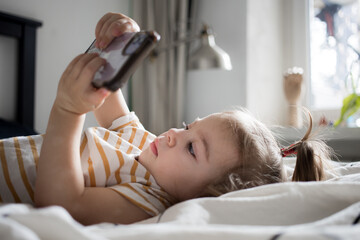 Obraz na płótnie Canvas Little toddle girl playing with smartphone laying on the bed at home. Caucasian toddler girl.