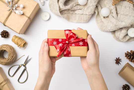 First person top view photo of woman's hands holding craft paper giftbox with red ribbon bow over scarf christmas tree balls and handicraft tools on isolated white background