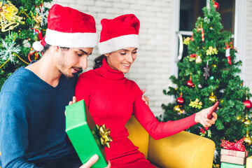 Romantic sweet couple in santa hats having fun decorating christmas tree and smiling while celebrating new year eve and enjoying spending time together.man giving gift box surprise to woman