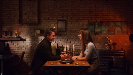 Elegant couple having romantic dinner at home, sitting at table in living room drinking red wine, talking. Stay at home concept.