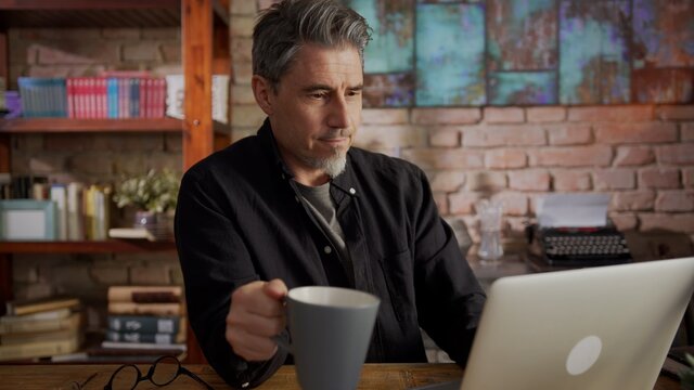 Mature man working from home, sitting at desk in home office using laptop, thinking, browsing internet, teleworking online.