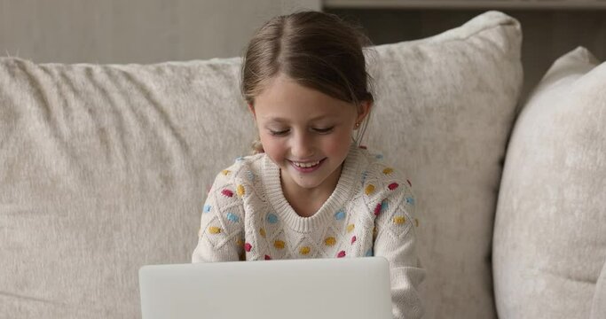 Modern tech addiction. Concentrated little girl sit on couch at home alone engaged in playing video game on laptop computer. Focused preteen child use pc device enjoy videogames need parental control