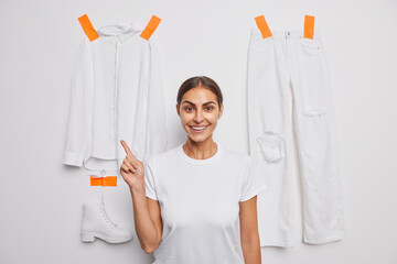 Waist up shot of good looking woman with dark hair indicates at items of clothes dressed casually poses against white background with plastered t shirt trousers and boot has glad expression.