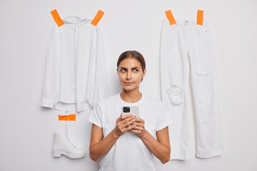 Thoughtful brunette woman dressed in casual t shirt uses mobile phone for making shopping online sells items of clothes in internet poses against white background. People and clothing concept
