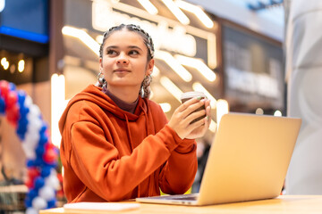 Alternative girl with white braids with a computer in a shopping center, pensive with a hot coffee in her hands