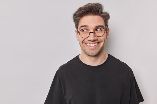 Portrait of handsome happy man looks curious aside wears round spectacles and casual black t shirt being in good mood isolated over white background with blank copy space area for your text.