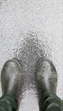 Close up, zoomed top-down view of rubber boots. A man standing in dark green waterproof pants and water-resistant boots. Waist view shot. Grey and wet asphalt with a texture.