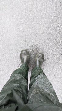 Top-down view of rubber boots. A man standing in dark green waterproof pants and water-resistant boots. Waist view shot. Grey and wet asphalt with a texture.