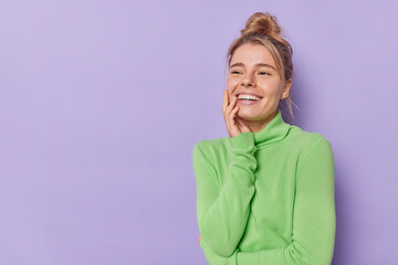 Horizontal shot of happy young woman smiles toothily looks somewhere feels glad thinks about something pleasant wears casual green turtleneck isolated over purple background with copy space.
