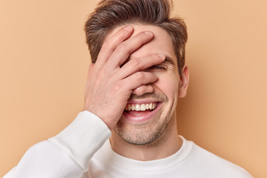 Close up shot of optimistic guy covers face with hand laughs joyfully smiles broadly hears something funny dressed in casual white jumper isolated over beige studio background hides emotions