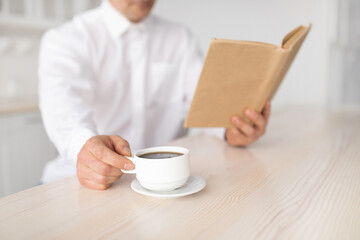 Unrecognizable mature man drinking coffee and reading book, enjoying peaceful morning at home, selective focus, crop