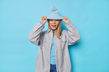 Positive young woman looks from under hat has fun feels glad dressed in grey jacket and jeans going to have walk isolated over blue background. Carefree sincere teenage girl wears outerwear.