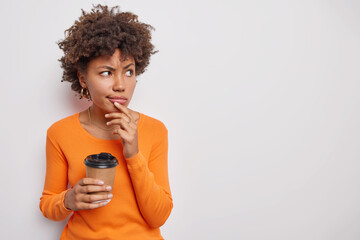Serious displeased young woman with curly hair looks attentively away drinks takeaway coffee thinks about something keeps hand on chin dressed in orange jumper isolated over white background