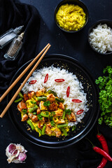 Fried chicken nuggets in curry sauce with white rice, green beans, parsley and chili peppers on black wooden table
