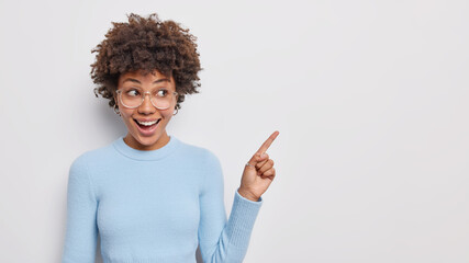 Place for your logo. Positive good looking young curly woman shows copy space points index finger at upper right corner demonstrates good product wears blue jumper isolated over white background.