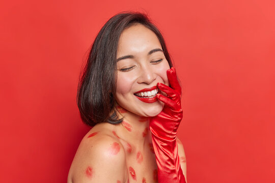 Portrait of brunette woman has lips traces on body stands with bare shoulders keeps eyes closed smiles happily shows white teeth wears long glove isolated over vivid red background glad to be in love