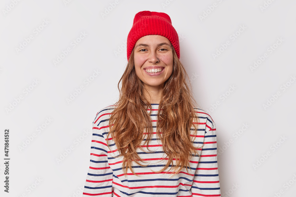 Wall mural Portrait of good looking young woman smiles happily looks joyfully has friendly conversation with friend wears red hat casual striped jumper isolated over white background. Happy emotions concept