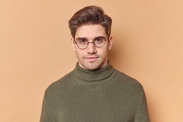 Fototapeta premium Portrait of handsome young European man looks directly at camera with serious expression feels self confident wears round transparent glasses and casual sweater isolated over beige background.