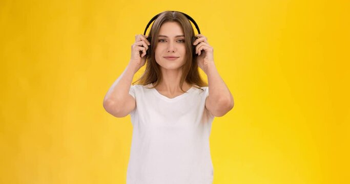 Dynamic music concept. Young cheerful woman putting headphones on and start dancing over yellow studio background