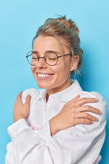 Pleased sincere woman embraces herself keeps arms crossed hands on shoulders feels cozy recalls lovely moment wears comfortable white shirt and spectacles isolated over blue background wraps body