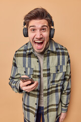 Emotional crazy man exclaims loudly listens music via wireless headphones holds mobile phone dressed in casual checkered shirt uses modern gadgets for listening music isolated over beige background
