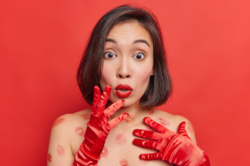 Surprised shocked brunette Asian woman gazes shocked at camera wears minimal makeup gasps from wonder stands with bare shoulders has lipstick traces on body isolated over vivid red background