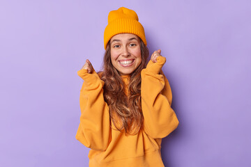 Naklejka premium Ecstatic positive woman clenches fists awaits for important results smiles broadly has white teeth feels excited feels enthusiastic and upbeat wears casual loose jumper and hat poses indoor.