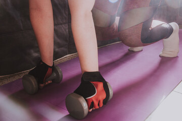 Detalle de una mujer haciendo ejercicio en casa sobre una esterilla de yoga púrpura. Mujer vistiendo guantes deportivos sosteniendo unas pequeñas mancuernas mientras hace una plancha - obrazy, fototapety, plakaty